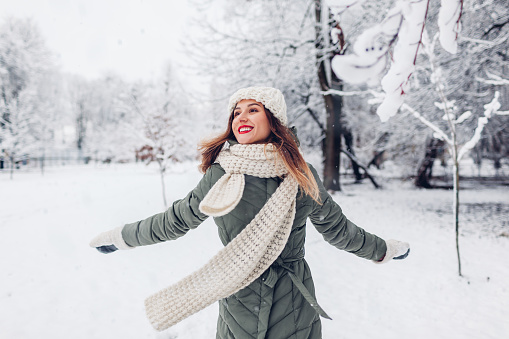Happy young woman spinning in snowy winter park wearing warm knitted clothes and having fun outdoors. Girl enjoying winter. Seasonal activities