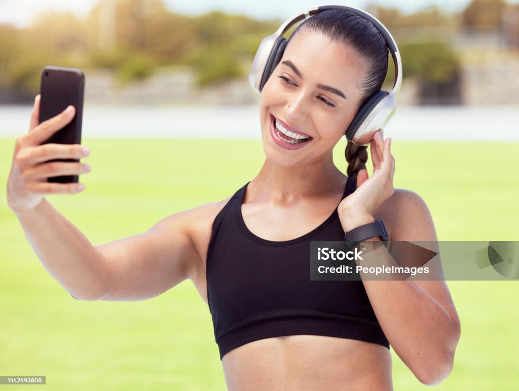 Phone, woman and fitness influencer taking a selfie at training, running and workout outdoors on sports racetrack. Smile, healthy and happy girl in headphones sharing fitness journey on social media 20-24 Years Stock Photo