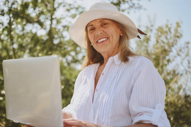 Senior happy woman working or having video call with laptop in summer outdoors. Old elegant lady in straw hat and bag sitting on grass during picnic at countryside. Active retired people concept. stock photo