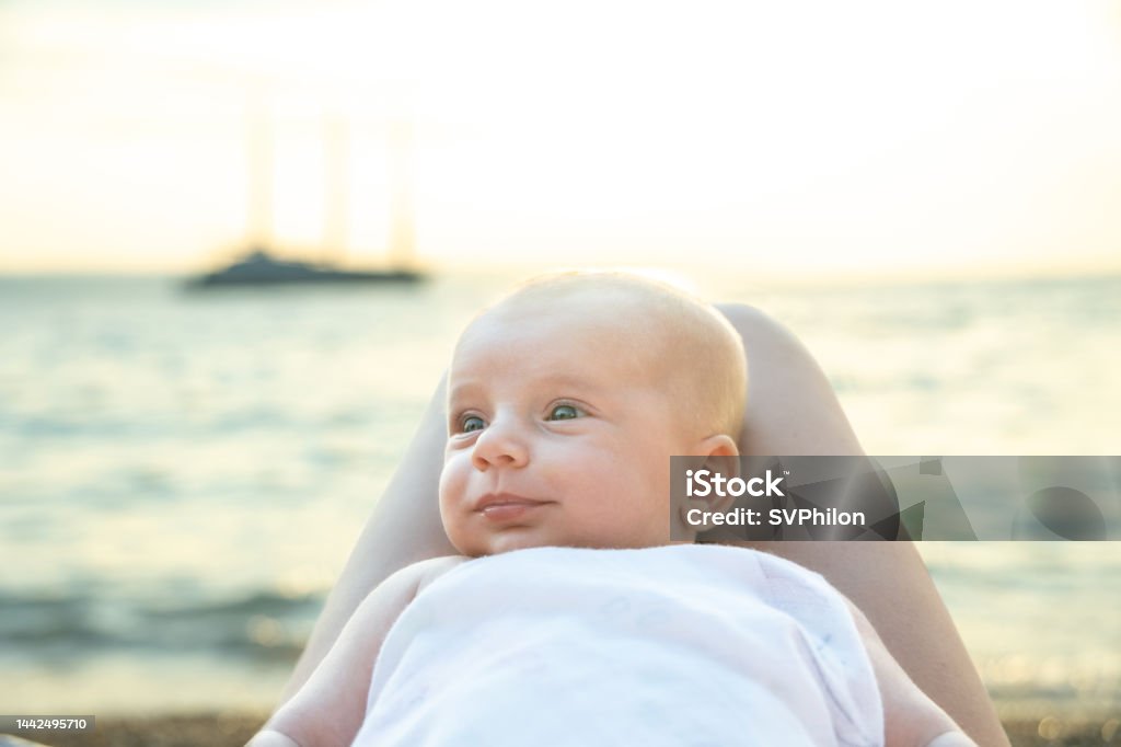 The baby lies on the legs of the mother near the sea in the evening. The baby lies on the mother's lap. Portrait of a baby boy against the background of an evening seascape. 
 Sunlight reflects on the surface of the sea water. 0-11 Months Stock Photo