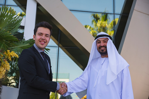 Young entrepreneur western business man with Emirati Arab sheikh man looking at camera and smiling while about presenting and shake hand struck a deal a new business projects and plans with digital tablet to poised and successful on behind of modern office outside. UAE