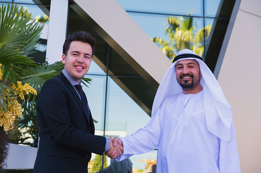Young entrepreneur western business man with Emirati Arab sheikh man looking at camera and smiling while about presenting and shake hand struck a deal a new business projects and plans with digital tablet to poised and successful on behind of modern office outside. UAE