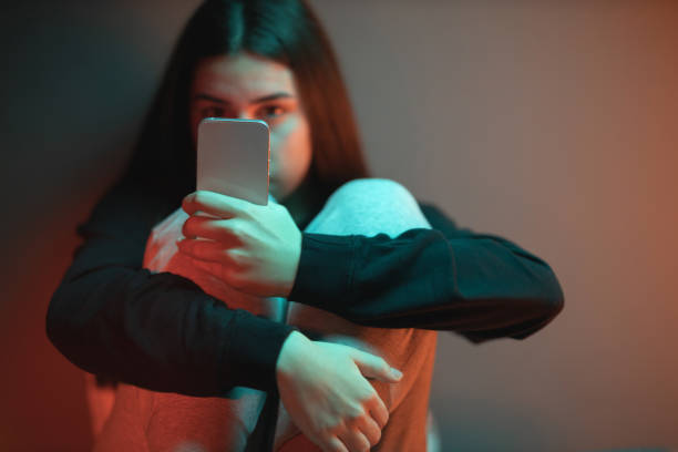 The Social Media Overuse Close-up of a depressed teenage girl sitting on the bed in her room lightened with orange and teal neon lights and scrolling social media. cyberbullying stock pictures, royalty-free photos & images