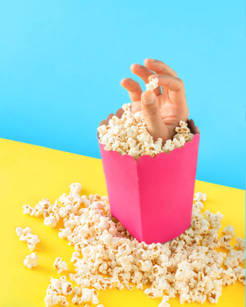 istock Pile of popcorn around a bold pink colored popcorn box with a man's hand holding a popcorn on isolated vibrant blue and yellow background. Surrealist abstract takeaway food concept. Pop art style. 1442506039