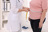 Nutritionist measuring overweight woman's waist with tape in clinic, closeup