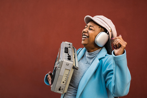 Happy trendy senior African woman having fun dancing while listening music with headphones and vintage boombox stereo