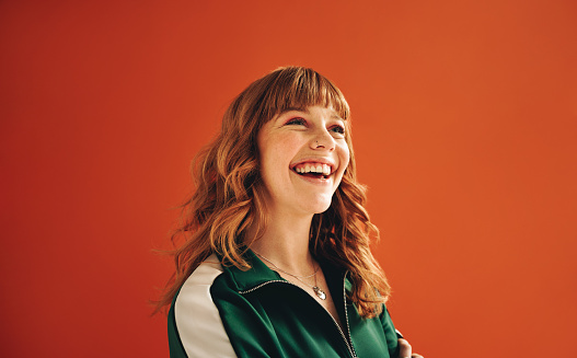 Happy young woman looking away with a smile while standing in a studio. Cheerful woman with ginger hair standing against a vibrant orange background in casual clothing.