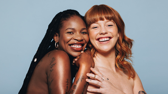 Happy women with different skin tones smiling at the camera while standing together. Two body confident young women embracing their natural beauty. Cheerful young women standing topless in a studio.