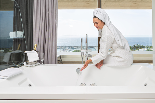 Side view of delighted woman in bathrobe and towel enjoying warm water from tap in modern hotel bathroom