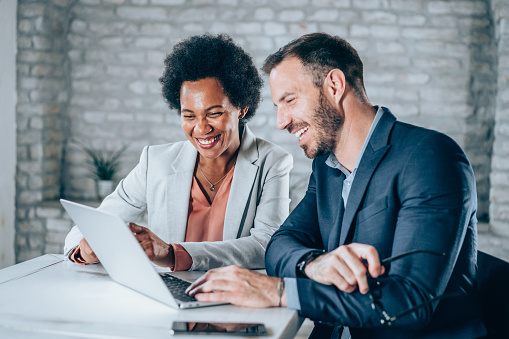 Shot of a two confident business persons sitting on a desk in the office. Businessman and businesswoman in meeting using laptop and discussing business strategy. Business coworkers working together in the office.