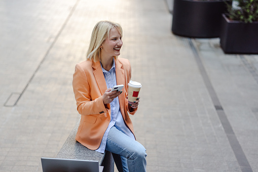 Blonde hair young business woman taking a coffee break and using smart phone in front of the office