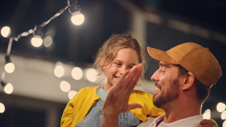 Cinematic Shot of a Father Holding His Small Daughter in His Arms, Helping Her to Fix a Lightbulb in a Backyard Lights Installation. Father and Daughter High Five and Celebrate Successful Repair.