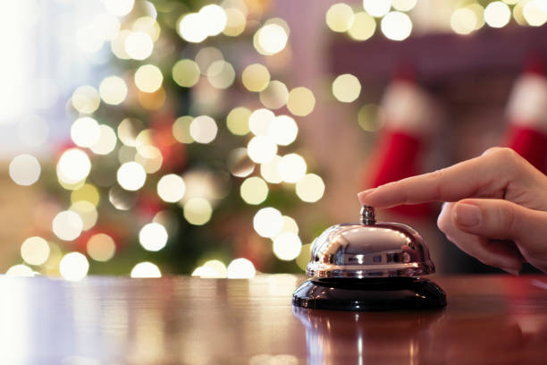 Hand of guest ringing reception bell on desk of guesthouse, hotel at christmas time. Color shining garland on christmas tree on background. Travel concept. stock photo