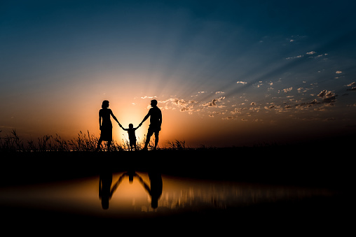 Silhouettes of Happy family holding hands with reflection on backdrop of the sea, sun, clouds in blue and red colors.