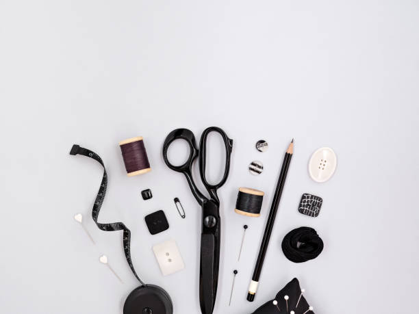 Assorted monochrome craft and sewing tools. Scissors, buttons, pins and measuring tape lying on grey background with copy space. Bottom frame, mock up for tailoring or dressmaking business design.