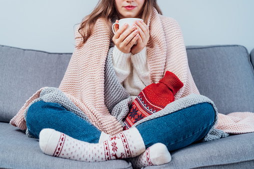 Woman freezes in wintertime. Young girl wearing warm woolen socks and wrapped into two blankets, holding a cup of hot drink and heating pad while sitting on sofa at home. Keep warm. Selective focus.