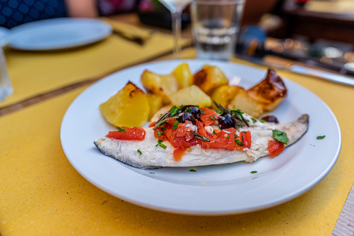 Fresh grilled fish with oven backed potatoes, juicy tomatoes, black olives, herbs and rosemary served on white plate with a glass of white wine, Italy, Europe