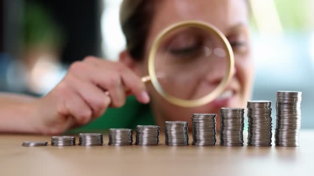 Businesswoman using magnifying glass examines stacks of coins in ascending order
