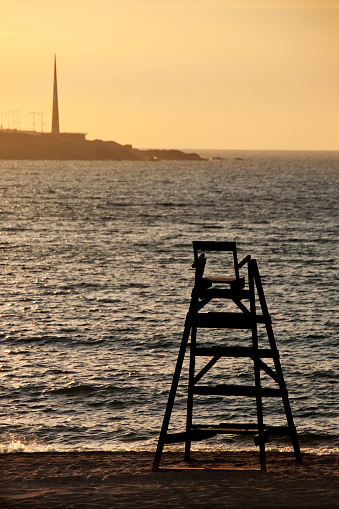 Lifeguard small wooden watchtower silhouetted, sunset seascape in the background, sunlight reflection on water surface. Millenium obelisque and coastline in La Coruña, Galicia, Spain.