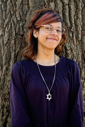 Transgender teenage girl portrait outdoors on a tree on a warm day in autumn. She multiracial, half latino, half european. She is also jewish, and is proudly wearing a star of david necklace. Vertical outdoors waist up shot with some copy space. This was taken in the suburbs of Montreal, Quebec, Canada.