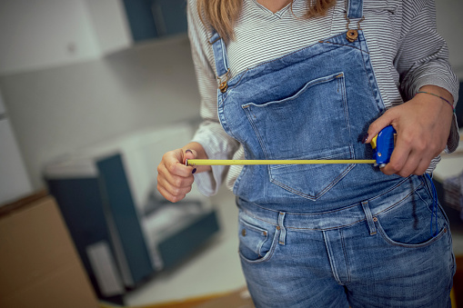 An unrecognizable woman uses a tape measure to measure the dimensions of kitchen elements during renovation