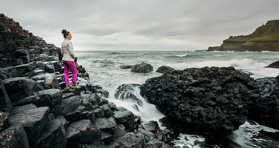 Young woman looking out at the rough ocean during a hike along a rocky coastline on an overcast day