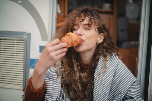 Front view of a woman sitting in front of a camper eating a croissant