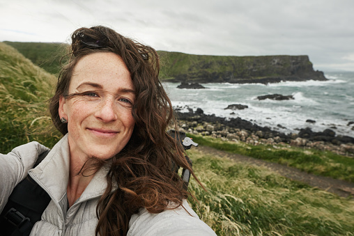 Smiling young woman taking a selfie during a scenic hike along a rugged coastline by the ocean on an overcast day
