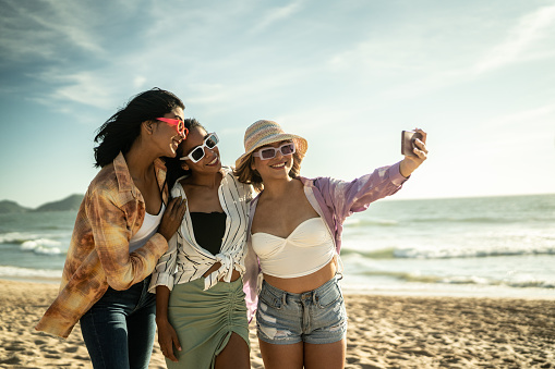 Young friends taking selfies or filming using mobile phone at the beach