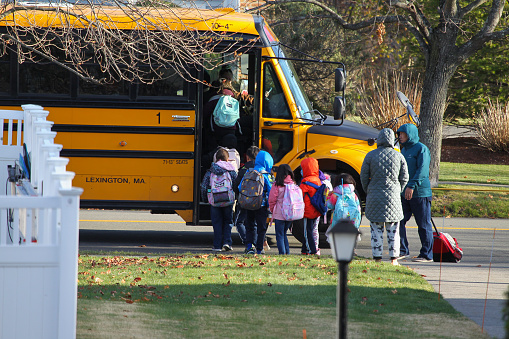 Elementary students waiting for school bus at Emerson Gardens Road,  Lexington, MA. November 18, 2022,\nMultiethnic mixed-race pupils classmates schoolchildren students standing in line waiting for boarding school bus in winter morning