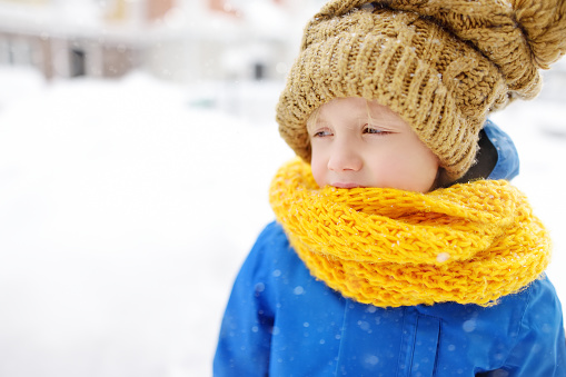 Upset little boy in blue winter clothes walks during a snowfall. Close-up portrait of crying school-age child. Real emotions. Sad and offended kid.