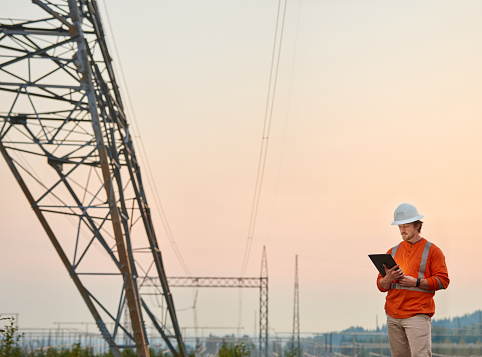 Male electrical engineer wearing reflective clothing and a hardhat standing with a digital tablet by power lines at dusk