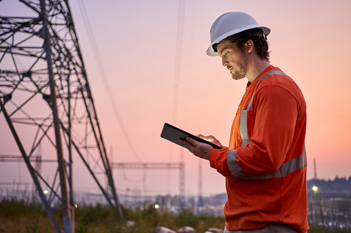 Young electrical engineer wearing reflective clothing and a hardhat using a digital tablet outdoors by power lines at dusk