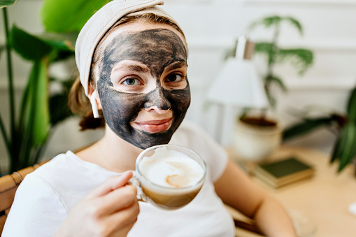 Woman with Charcoal Facial Cleansing Mask relaxing at home with cup of coffee