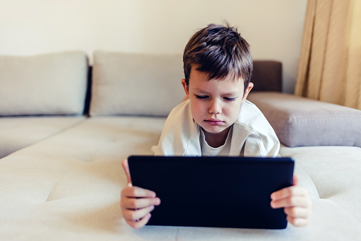 Boy lying in bed playing games on digital tablet at home. Little Caucasian boy sitting on sofa and playing game on digital tablet.