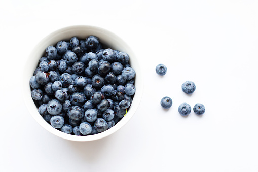 A white bowl full of blueberries stands on a white table and five berries lie nearby. View from above. Soft focus.