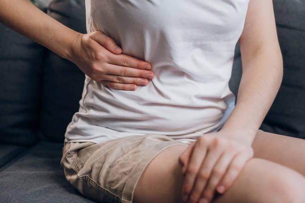 nahaufnahme einer jungen frau mit fettleber berührt die rechte seite mit der hand, die an bauchschmerzen leidet und auf einer grauen couch sitzt. schmerz in der rechten seite, blinddarm, gallensteine und gynäkologischen erkrankungen konzept - belly ache stock-fotos und bilder