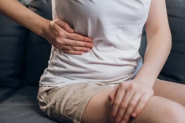 Photo of Close up of young female with fatty liver touches right side with hand, suffering from abdominal pain sit on grey couch. Pain in right side, appendix, gallstones and gynecological diseases concept