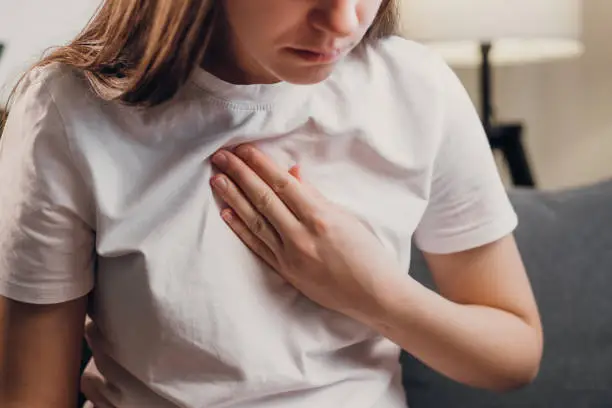 Photo of Young unhealthy female sit on couch having difficulty breathing pain of heart, touches his chest with hand. Trouble breathing, chest pain. Heart attack, thoracic osteochondrosis, panic attack concept