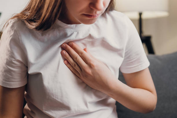 young unhealthy female sit on couch having difficulty breathing pain of heart, touches his chest with hand. trouble breathing, chest pain. heart attack, thoracic osteochondrosis, panic attack concept - peito imagens e fotografias de stock