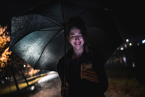 Image of a caucasian girl using the phone with an umbrella in her hand in the rain outside