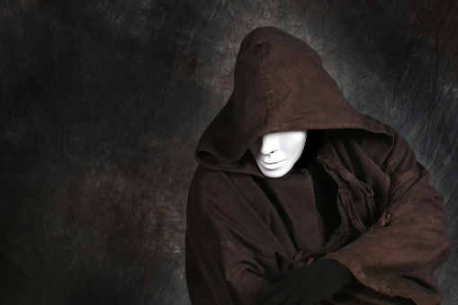 Horizontal photo of a faceless person (demon, monster) wearing a white theater mask, standing against a dark background, wearing a long hooded duster coat, and crossing his arms