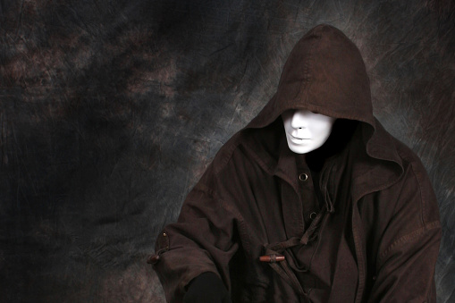 Horizontal photo of a faceless person (demon, monster) wearing a white theater mask, standing against a dark background, wearing a long hooded duster coat, and looking away.