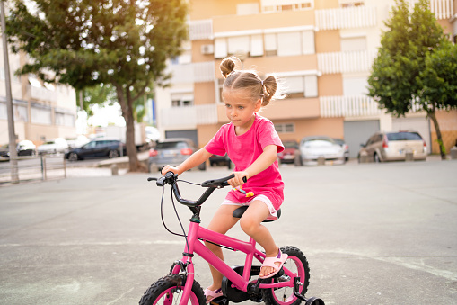 Cute little smiling girl riding bike bicycle in city on parking sunny summer day. Active family leisure with kids. Little caucasian girl 5 years old have fun with pink child bicycle