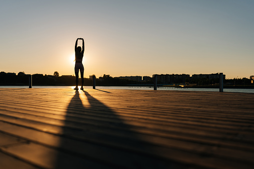 Silhouette of unrecognizable athletic woman in sporty outfit warm up muscles, practicing stretching exercises before running by water in city park. Sportswoman exercising in sunlight of summer morning