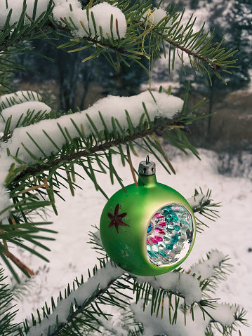 Close up of a traditional glass Christmas ornament hanging on the snow covered branch of an evergreen branch in a forest