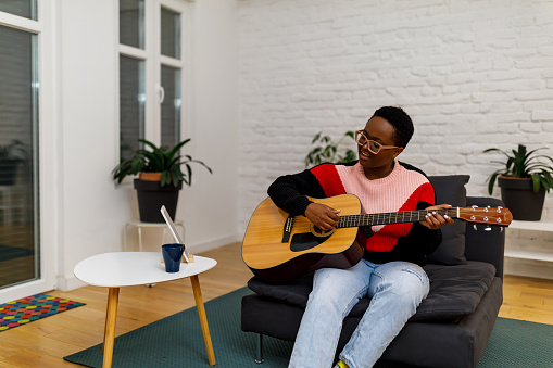 A young African woman is sitting on a sofa in the living room and playing an acoustic guitar and enjoying the sounds of the music.
