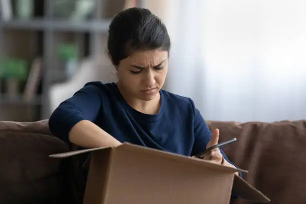 Stressed young Indian woman unpacking carton box, holding cellphone in hands. Unhappy female consumer dissatisfied with getting wrong or damaged order, negative internet shopping experience concept.