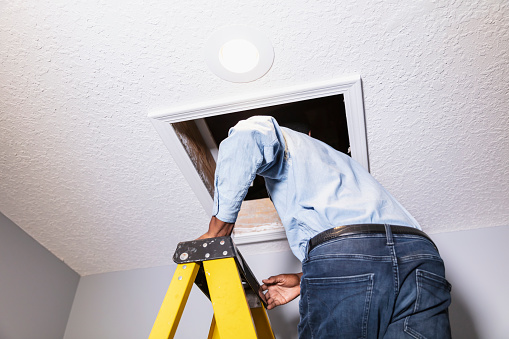 An African-American man on a ladder opening a door in the ceiling leading to the attic of a house. He is a house inspector or repairman or home owner.