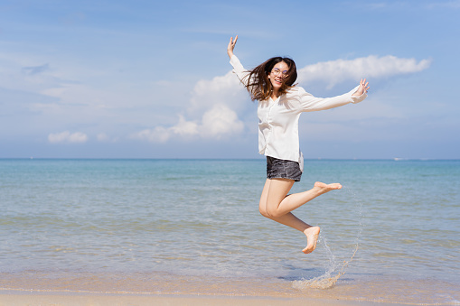 Happy Asian young woman enjoy playing and splashing a sea water at the beach. Summertime holiday and vacation concept. Cheerful Asian woman walking along the beach in morning and looking at camera.